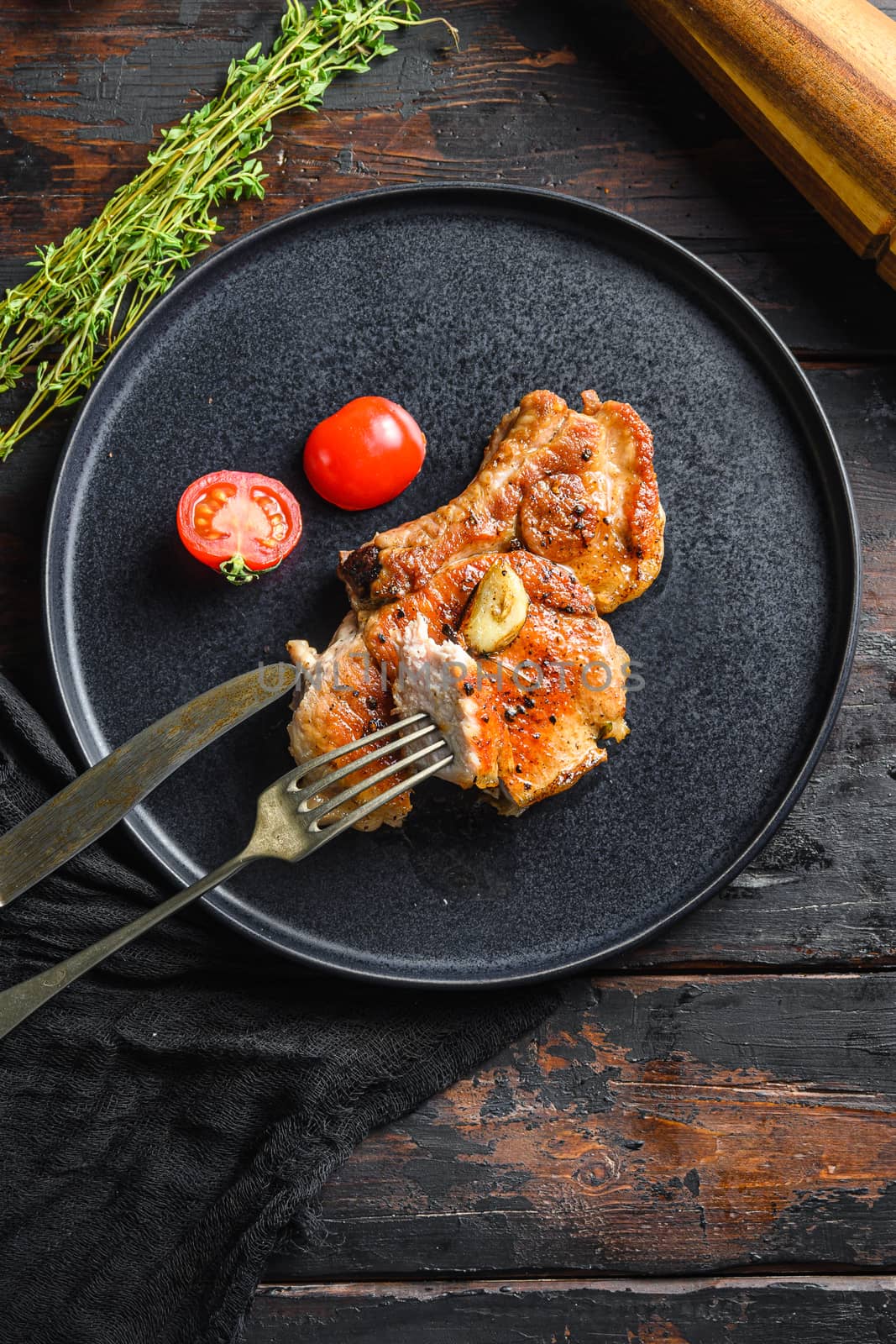 Dish of grilled pork chop with tomatoes top view with knife and slice on fork over old rustic dark wood table table flatlay top view vertical.