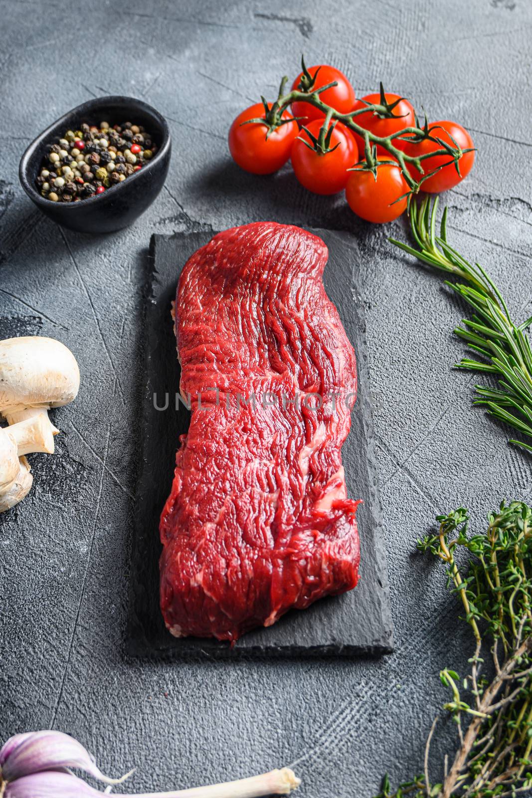 Organic Tri-tip, triangle roast marbled beef on black plate , marbled beef with herbs tomatoes peppercorns over grey stone surface background side view vertical.