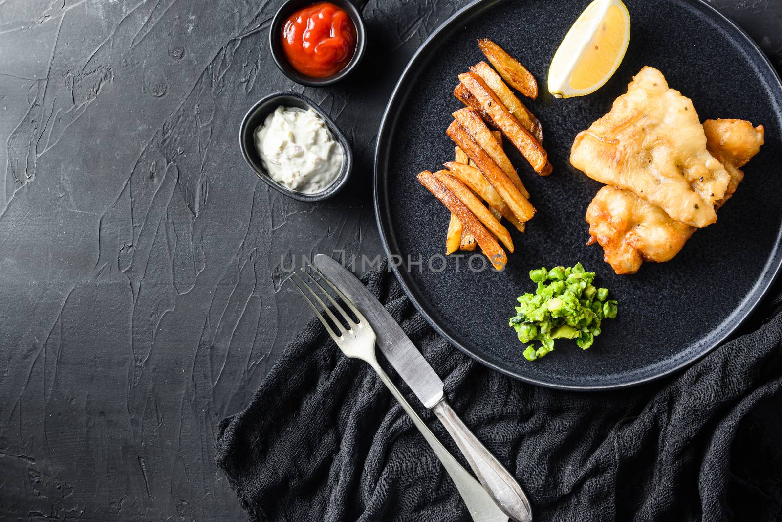 Fish chips with dip and lemon - fried cod, french fries, lemon slices, tartar sauce, ketchup tomatoe and mushy peas with fork and knife on black plate over black background top view space for text by Ilianesolenyi