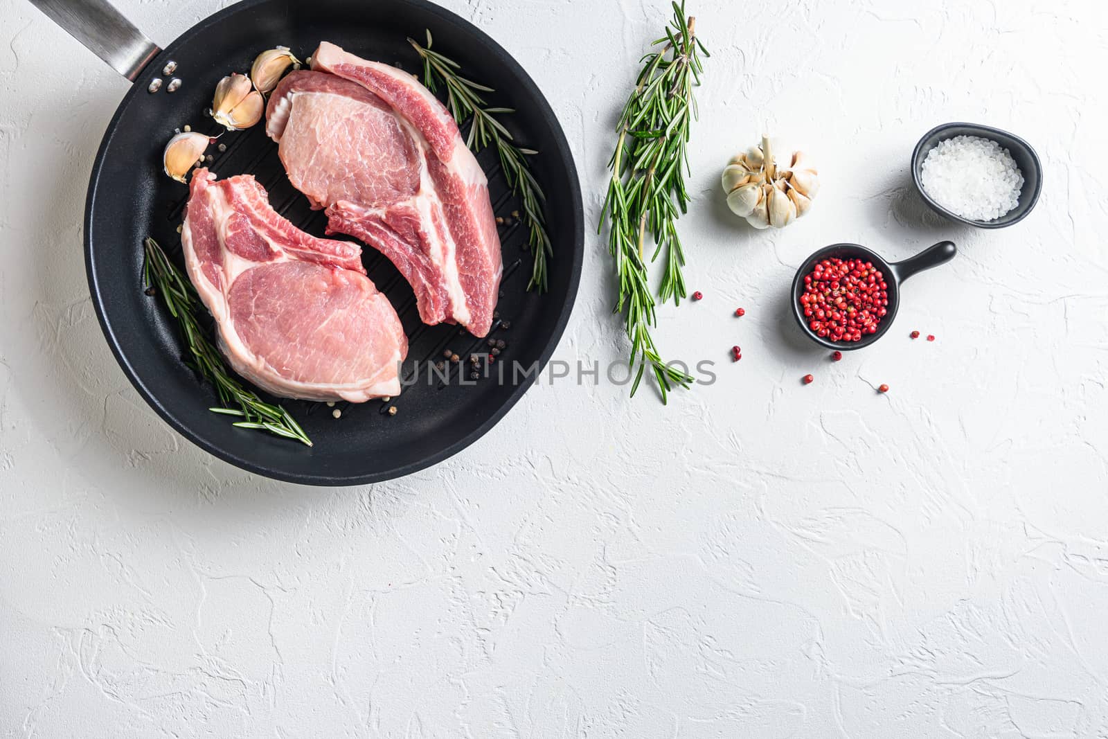 Fresh pork steak in a frying grill pann on a white stone surface with rosemary garlic peppercorns ingredients for grill top view side for text concept.