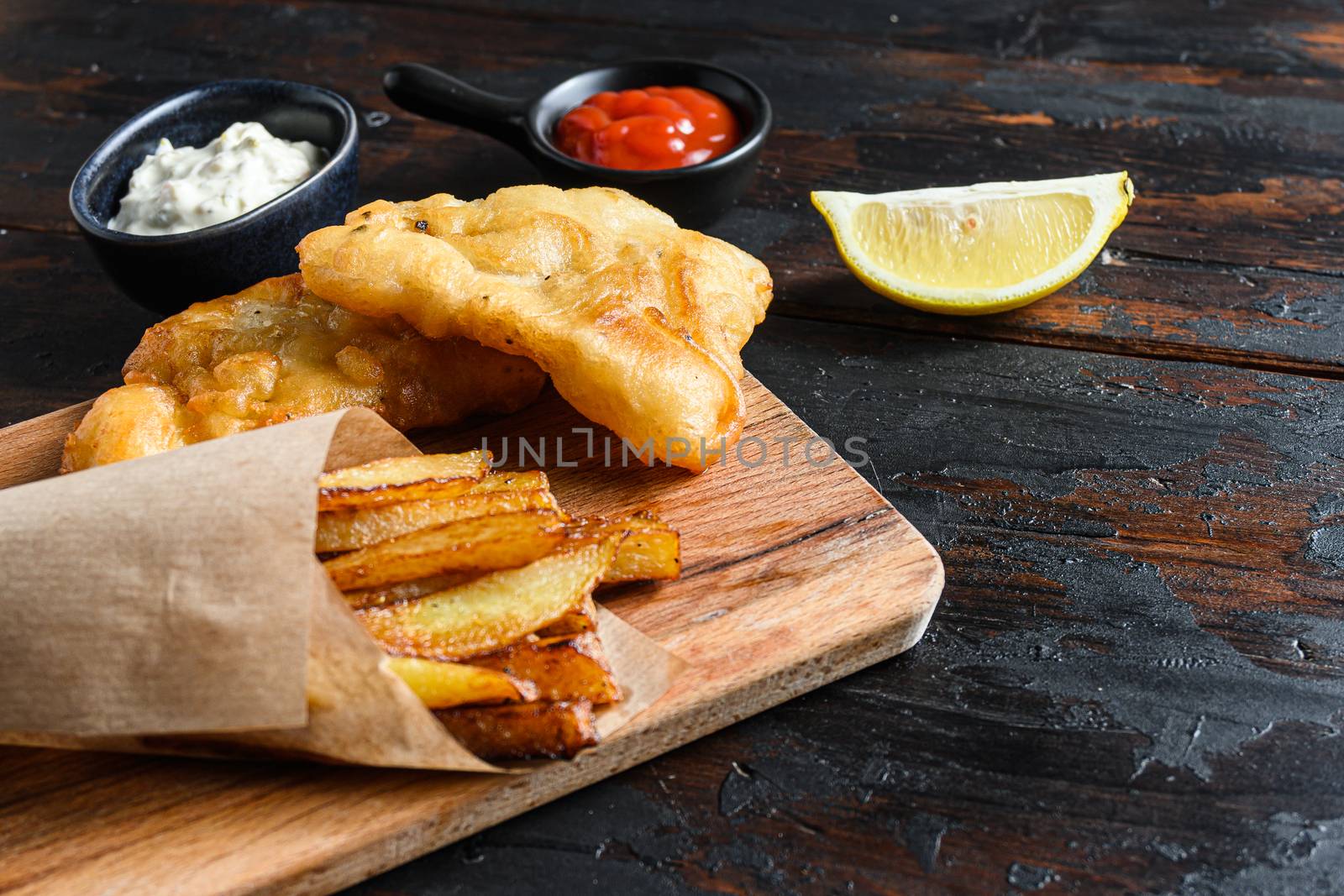 Fish and chips in a paper cone on wood chopping board dip and lemon - fried cod, french fries, lemon slices, tartar sauce, ketchup tomatoe and with mushy peas served in the Pub or Restaurant over old wooden planks dark table side view close up space for text.