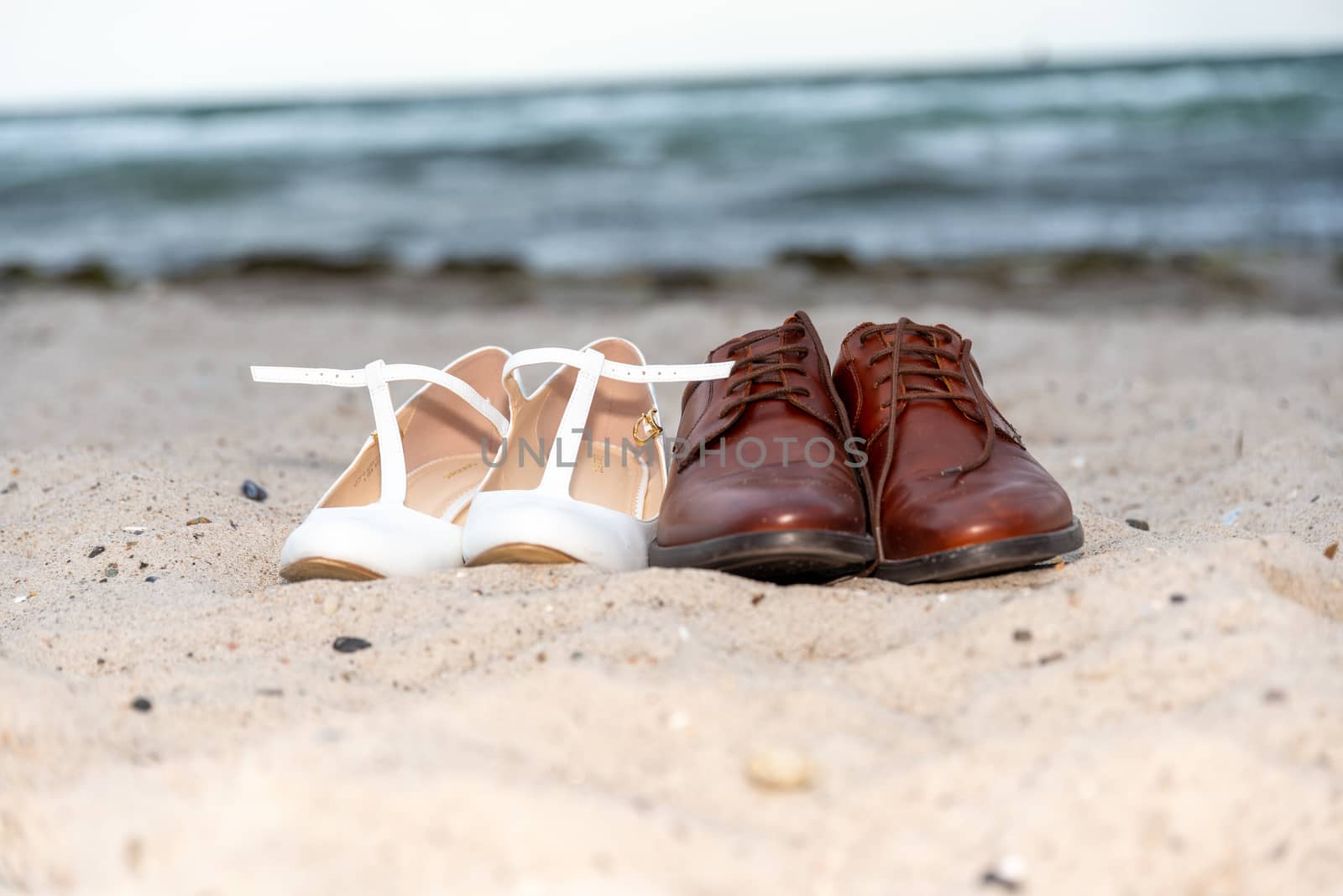 Fehmarn, Schleswig-Holstein/Germany - 03.09.2019: A white pair of ladies' shoes and a pair of brown men's shoes are standing on the beach in the sand with water in the background.