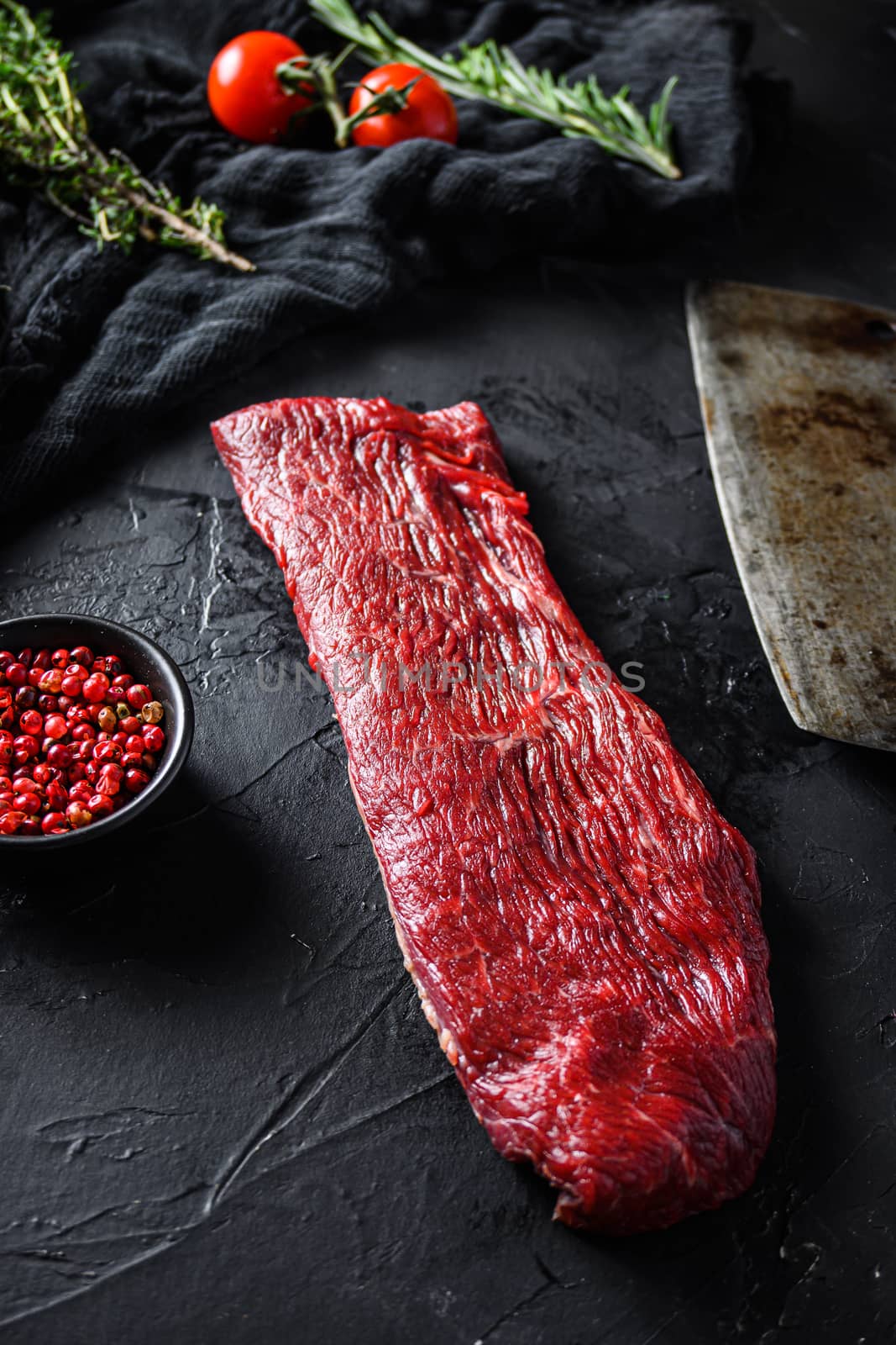 Raw triangle roast or tri tip, near butcher knife with pink pepper and rosemary. Black background. Close up side view selective focus.