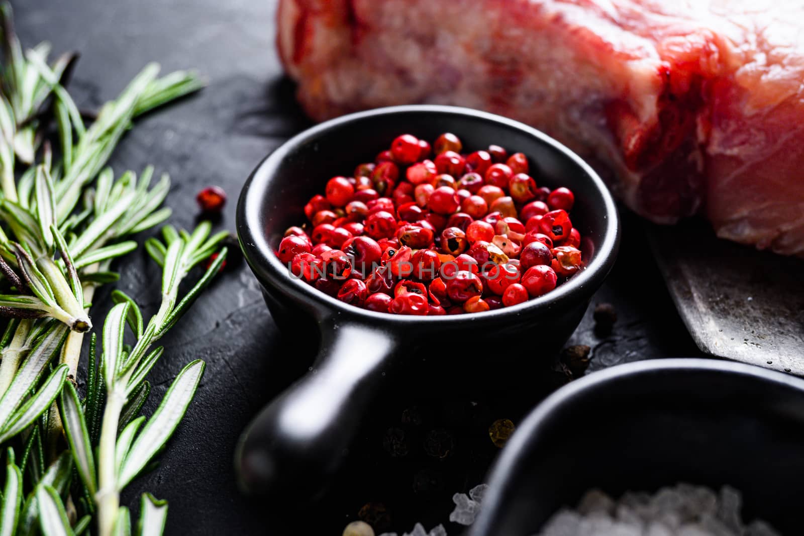 Rose Peppercorn and Rosemary herbs close up on black stone table with spices and raw meat nea rside view selective focus.