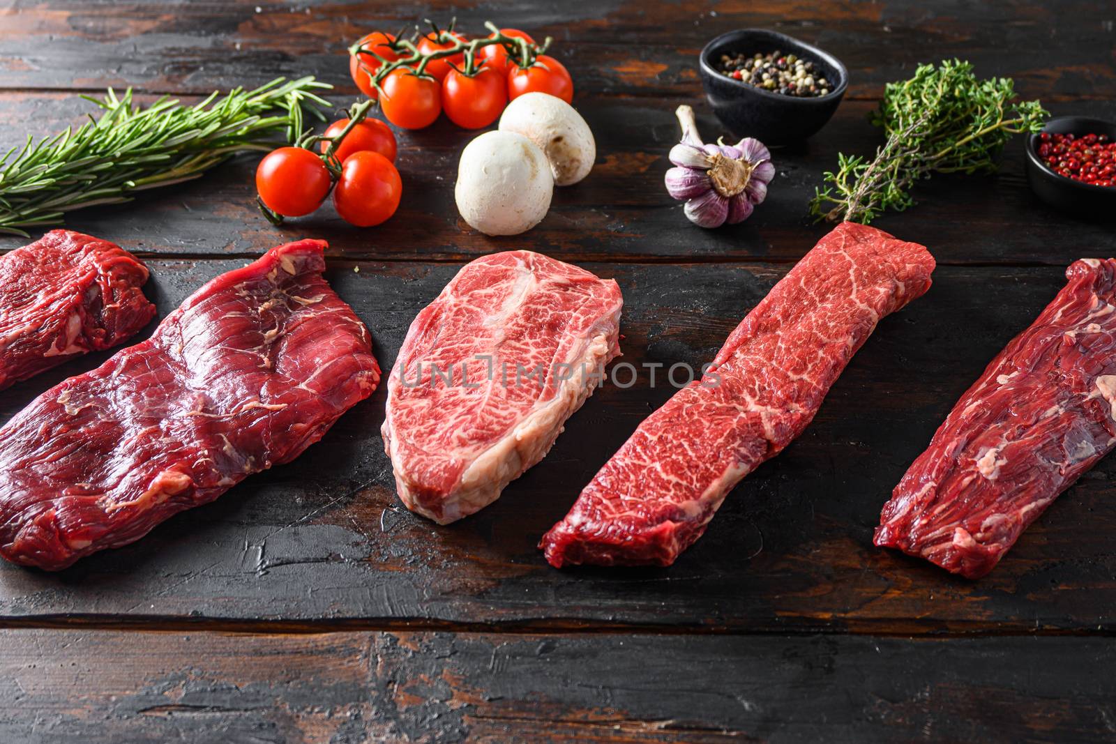 A set of different types of raw beef steaks alternative cut flap flank Steak, machete steak or skirt cut, Top blade or flat iron beef and tri tip, triangle roast with denver cut with fresh organic herbs over wood background side view space for text.