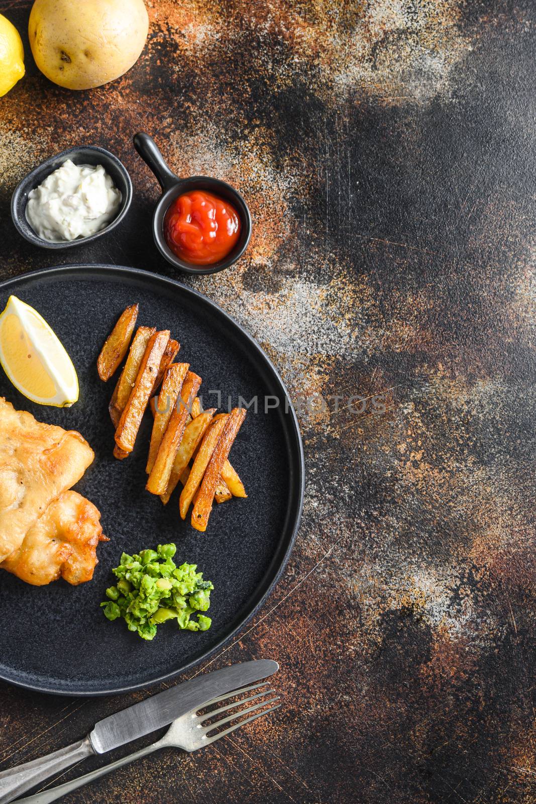 English Traditional Fish and chips with mashed minty peas, tartar sauce vertical side view on black plate Over old rustic table top view vertical concept with space for recipe or text.