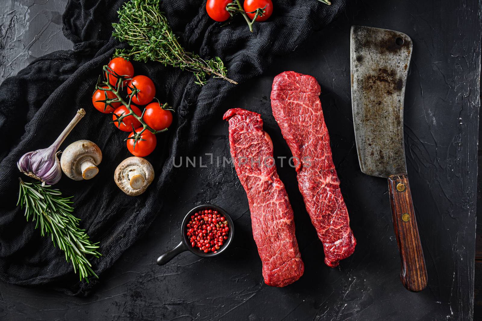 Raw set of denver cut organic meat with vegetable rosemary and other ingredients near butcher meat clever knife for bbq or grill over black stone background top view.