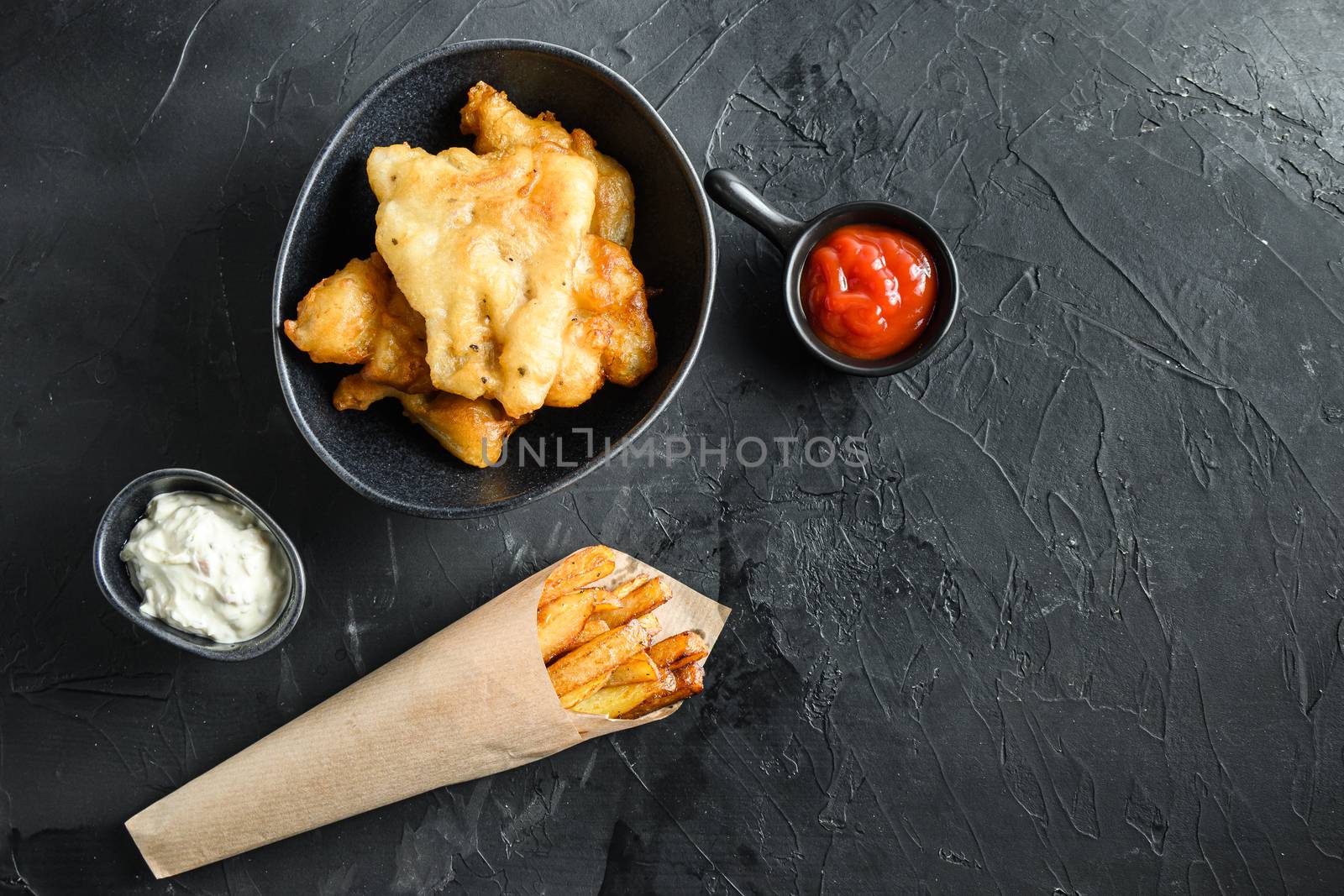 Fish and chips in a paper cone dip and lemon - fried cod, french fries, lemon slices, tartar sauce, ketchup tomatoe and mushy peas over black background top view space for text. by Ilianesolenyi