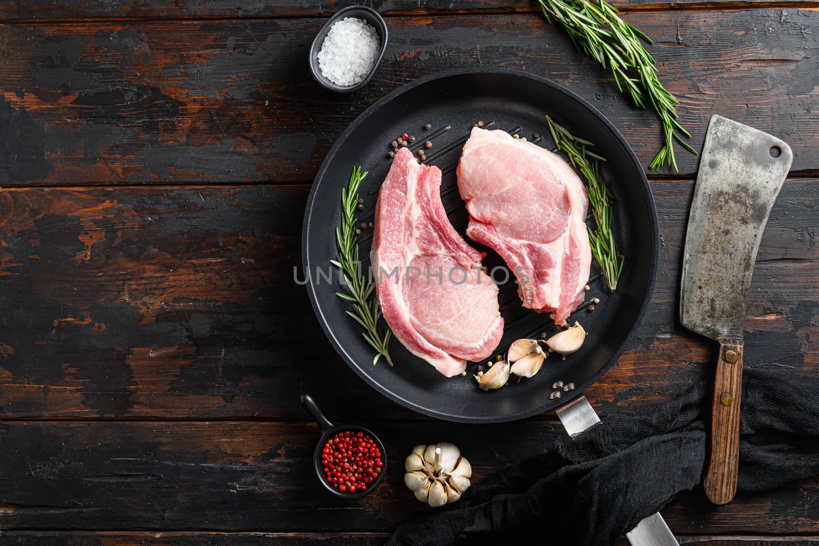 Raw pork meat chopes with herbs in iron skillet with burcher cleaver knife for chopping in butchery on a dark wood surface and ingredients for cooking. Food background with copy space. Top view by Ilianesolenyi
