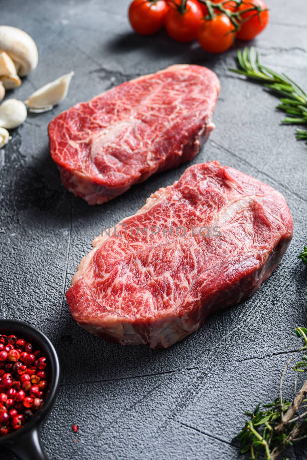 Raw top blade flat Ironcut, on black slate , bio marbled beef with herbs tomatoes peppercorns over grey stone surface background side vertical close up.