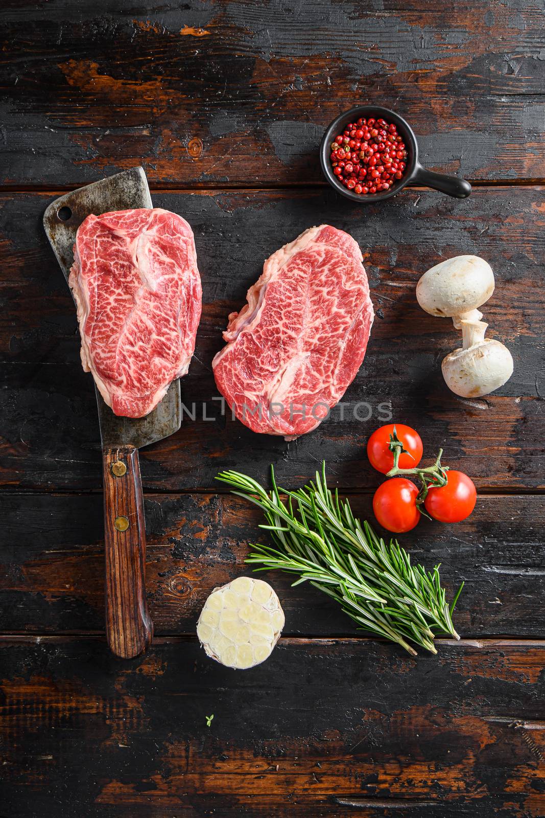 Organic Top blade steak, raw meat, marbled beef butler cut on metal butcher cleaver knife with rosemary and farm herbs over old wood table background top view.