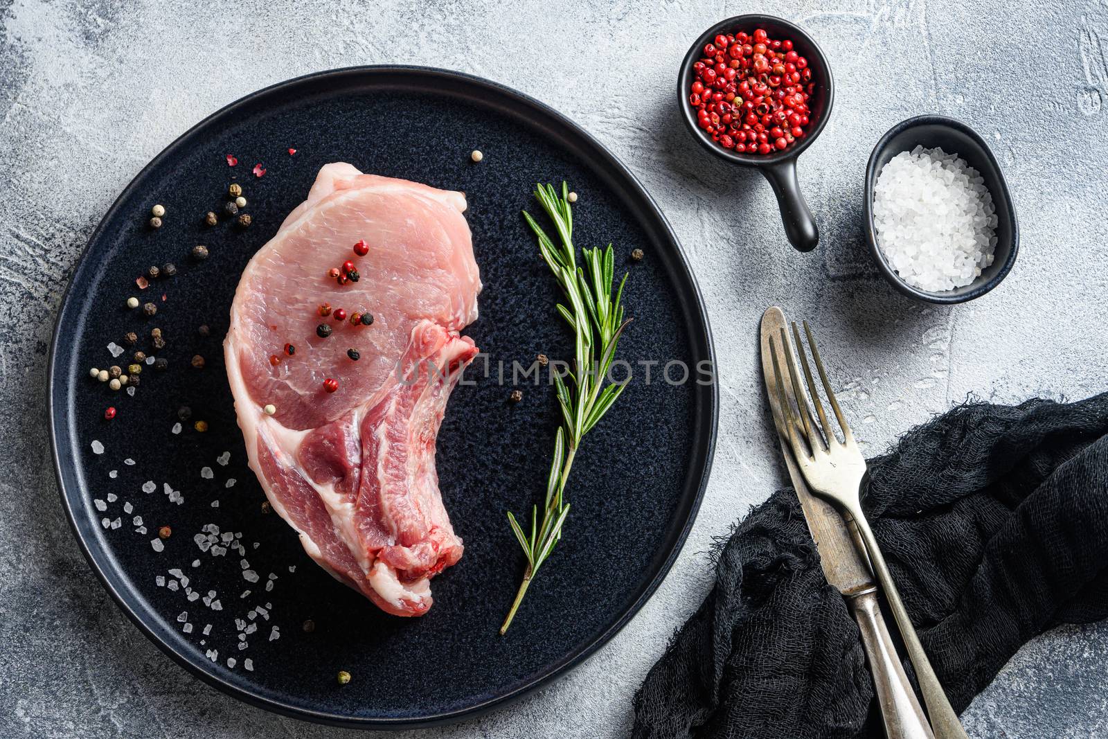 Raw Pork Loin chops in a black round plate on a grey textured stone background with rosemary garlic peppercorns ingredients for grill top view .