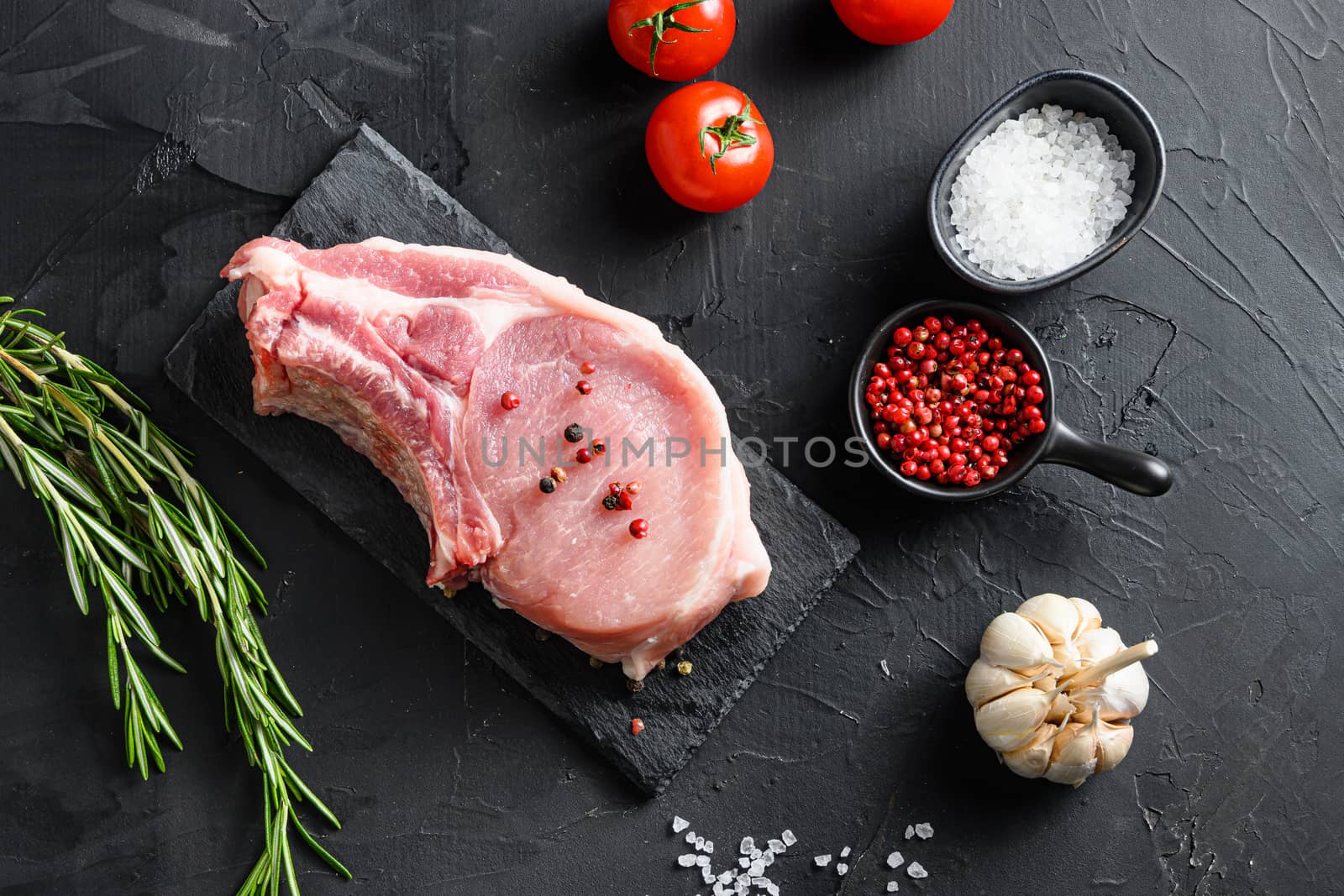 Top view raw pork chop steak and salt, pepper on black slate stone board with rosemary, and fresh ingredients for grill over black textured background.