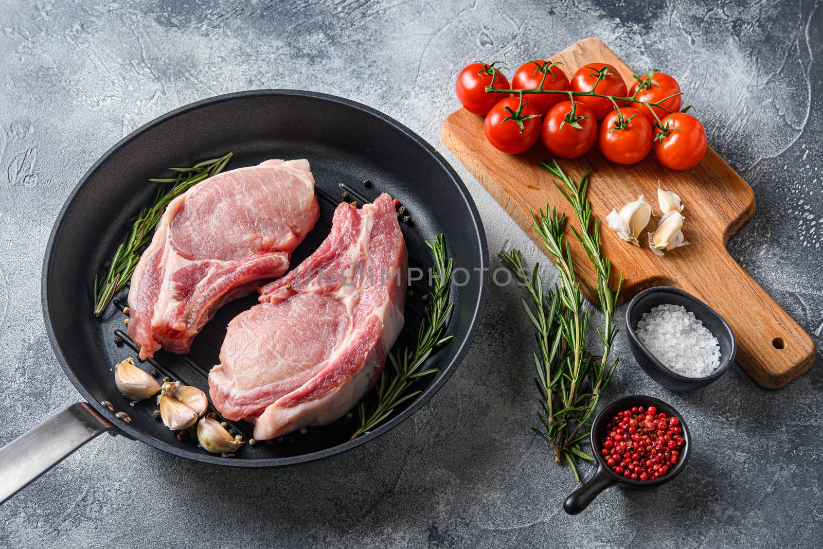 Pork chop on a bone in a black frying grill pan on a grey textured stone background with black cloth rosemary garlic peppercorns and tomatoes on chopping board ingredients side view .