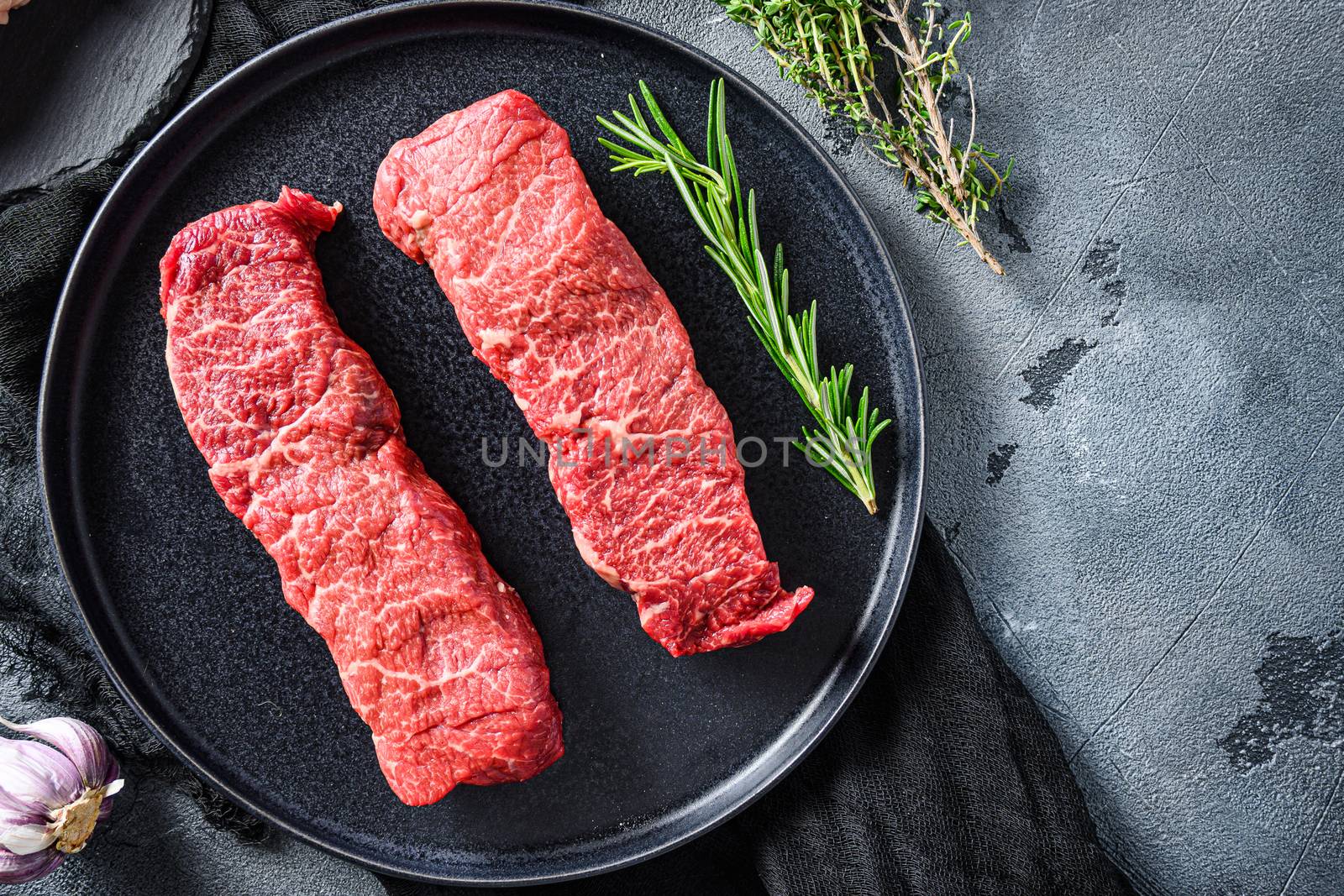 Raw denver cut black angus organic steak on a black plate and stone slate with seasonings, herbs grey concrete background. Top view space for text