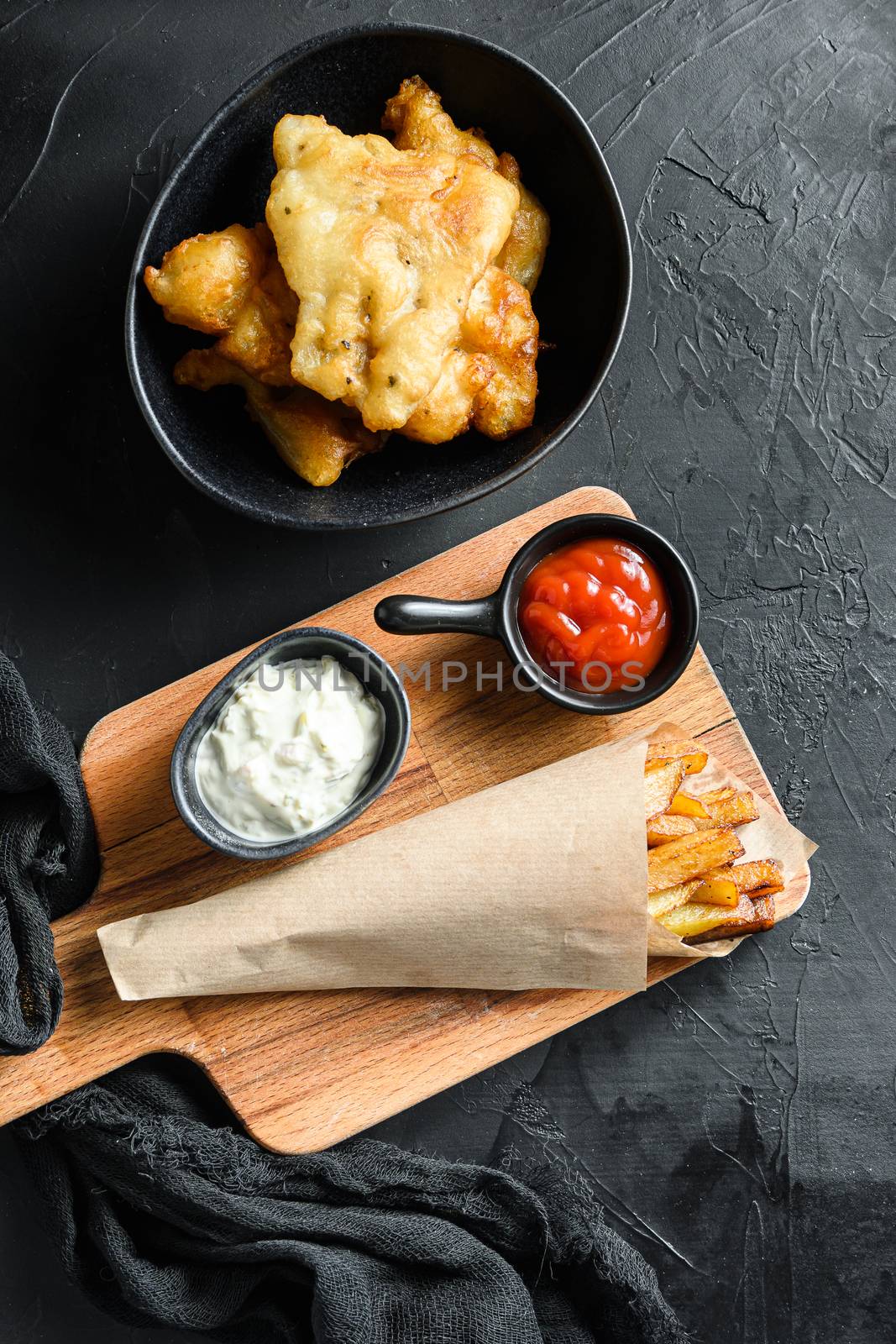 Fish and chips in a paper cone dip and lemon - fried cod, french fries, lemon slices, tartar sauce, ketchup tomatoe and mushy peas over black background top view vertical by Ilianesolenyi