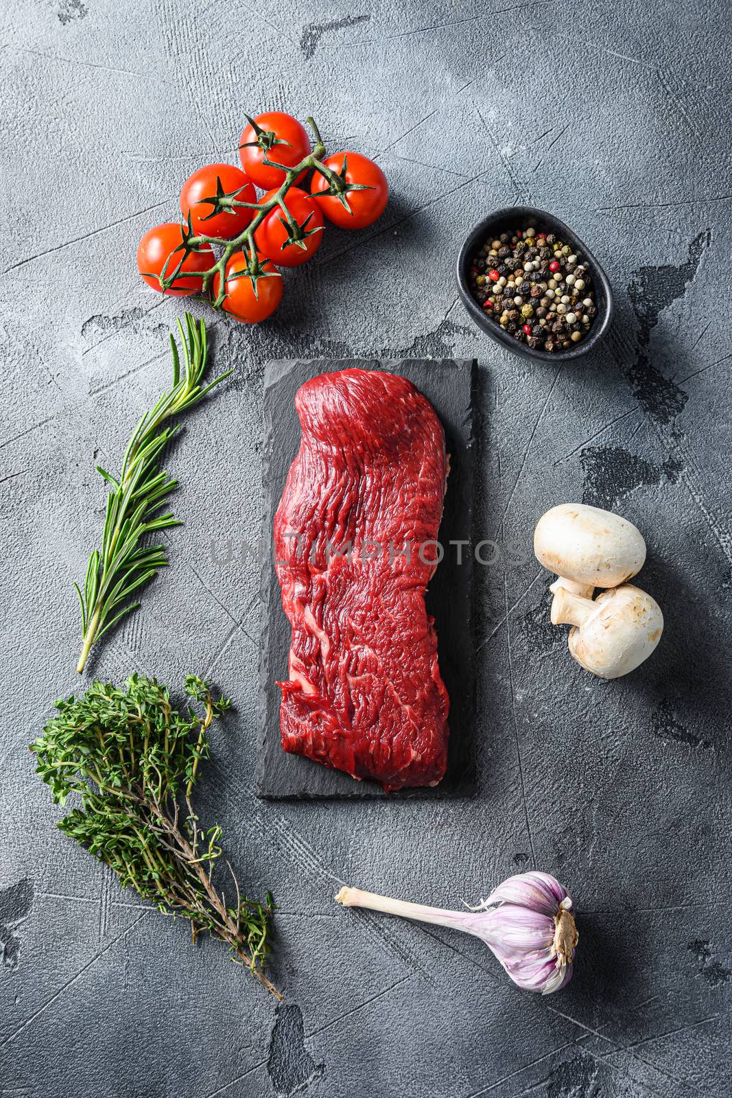 Organic Tri-tip, triangle roast marbled beef on black plate , marbled beef with herbs tomatoes peppercorns over grey stone surface background top view.