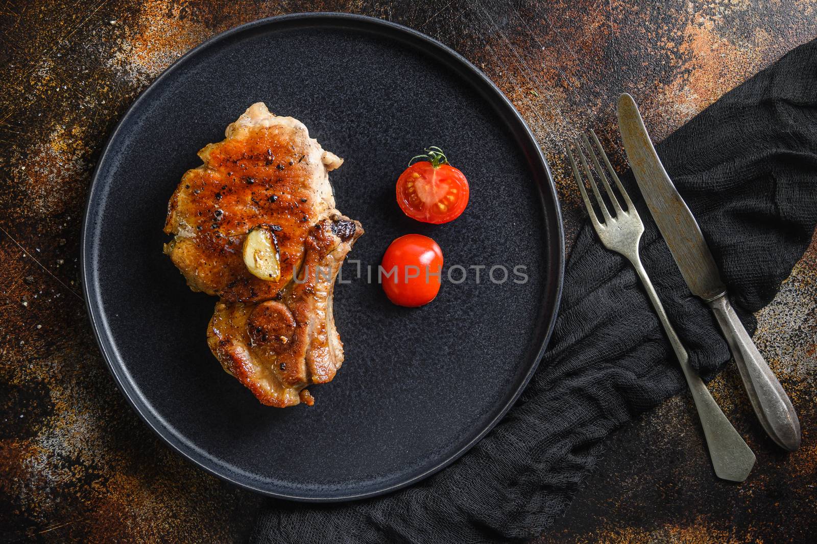 Grilled pork steak on the bone with seasonings on black dish with sfork and knife on cloth, over rustic metall surface overhead top view