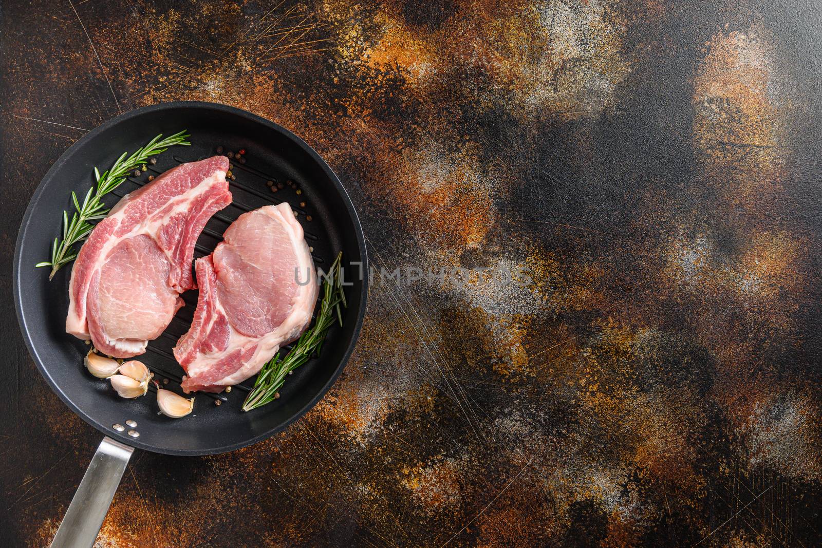 Raw Pork Loin chops on a bone in a black frying grill pan on a old rustic dark metall surface rosemary garlic peppercorns top view space for text.