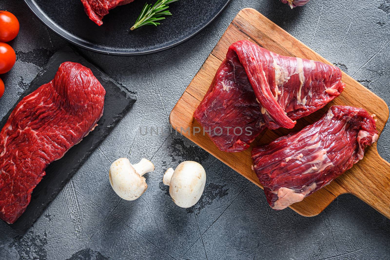 Organic Machete and flank, bavette steak on chopping board, marbled beef over other alternative cuts with herbs tomatoes peppercorns over grey stone surface background top view.