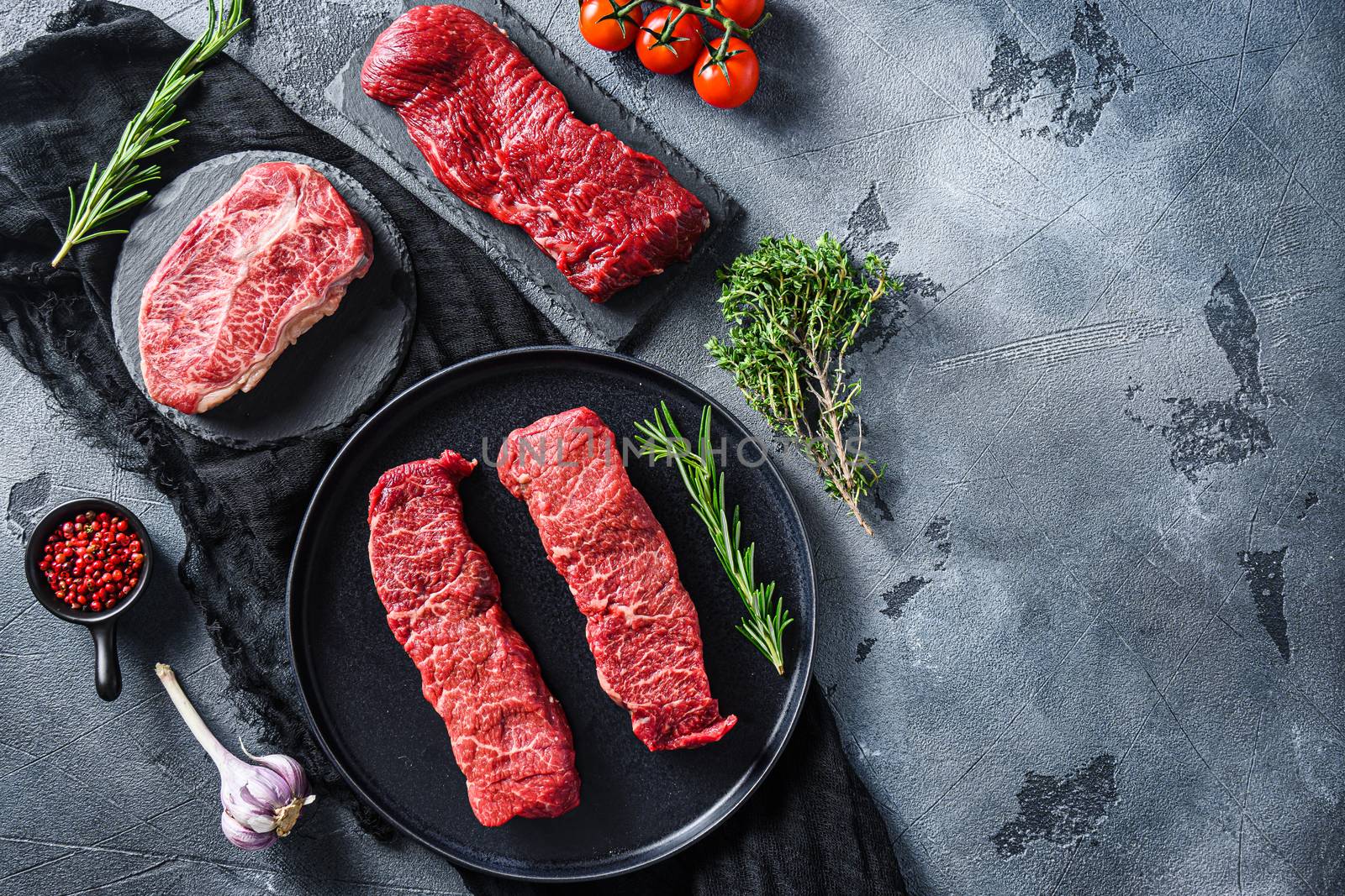 Raw denver, top blade, tri tip steak on a black plate and stone slate with seasonings, herbs grey concrete background. Top view space for text.