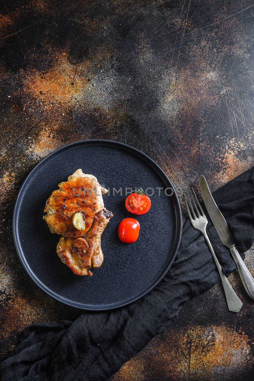 Grilled pork steak on the bone with seasonings on black dish with sfork and knife on cloth, over rustic metall surface overhead top view vertical space for text on top.