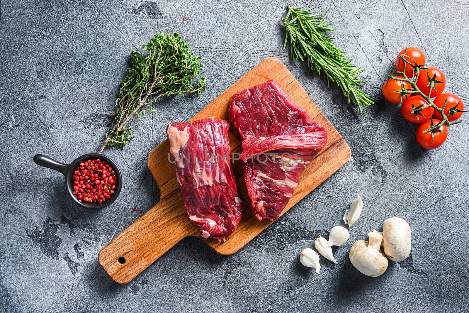 Raw flap steak flank cut with Machete, Skirt Steak, on woods chopping board, with herbs tomatoes peppercorns over grey stone surface background top view .