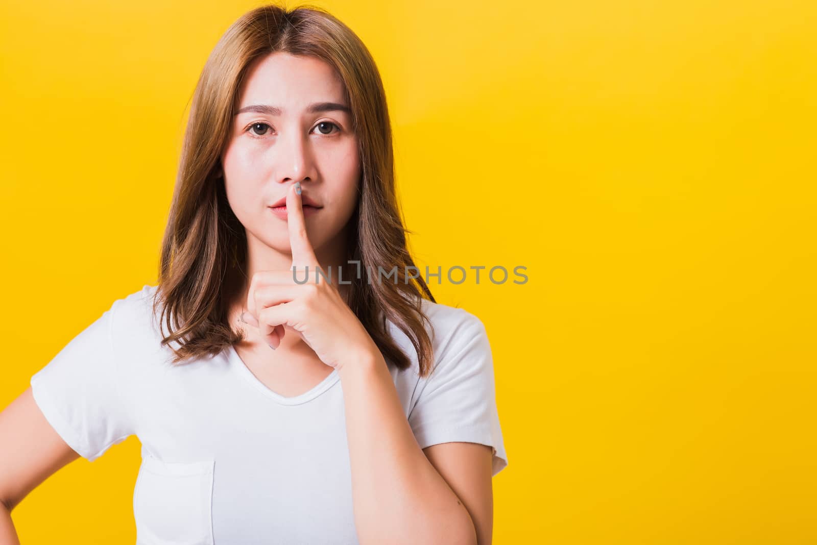 Asian Thai happy portrait beautiful cute young woman standing wear white t-shirt making finger on lips silent quiet gesture looking to side, studio shot isolated on yellow background with copy space