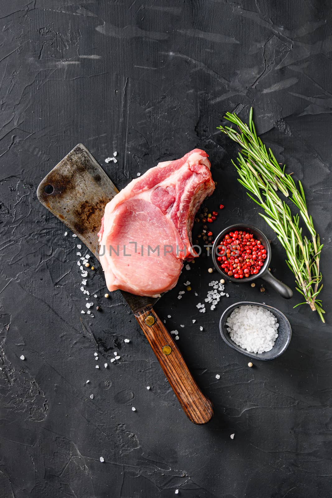 Organic cutlet on a rib or Pork meat over american classic butcher knife or cleaver with spices and rosemary and red pepper on black slate top view. vertical by Ilianesolenyi