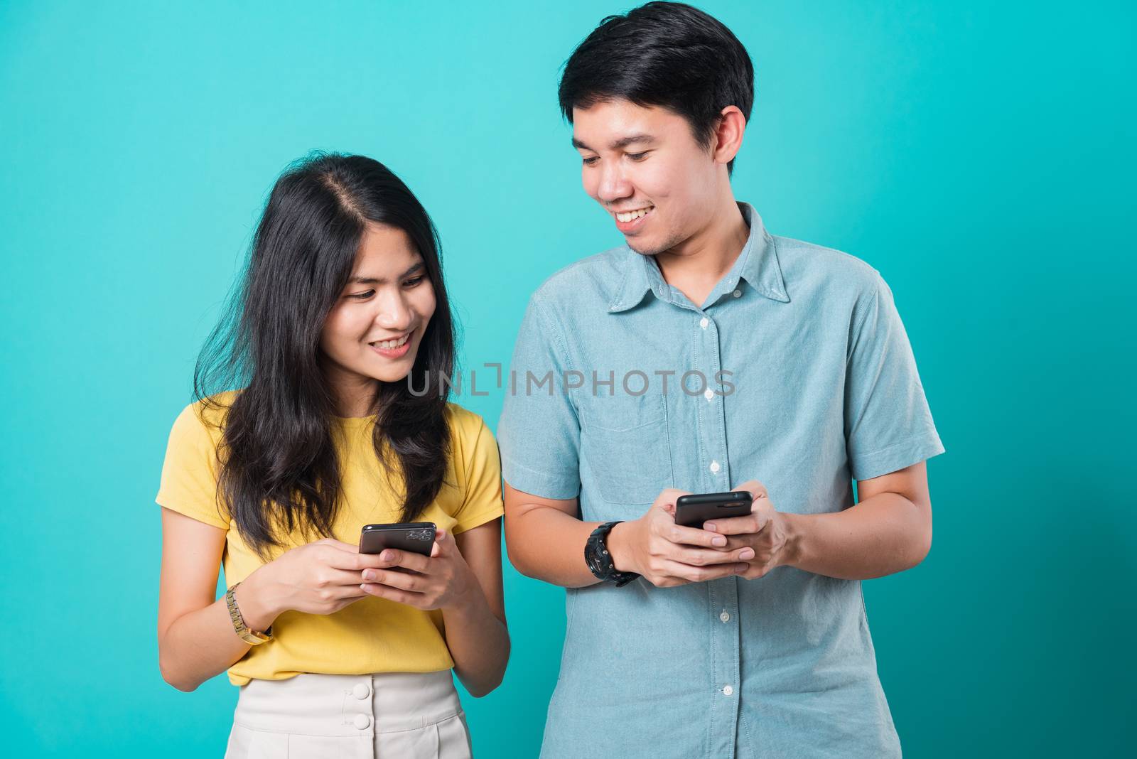 Portrait happy Asian young handsome man, beautiful woman couple smile standing wear shirt, excited young couple holding mobile phones looking at cellphone, studio shot on blue background