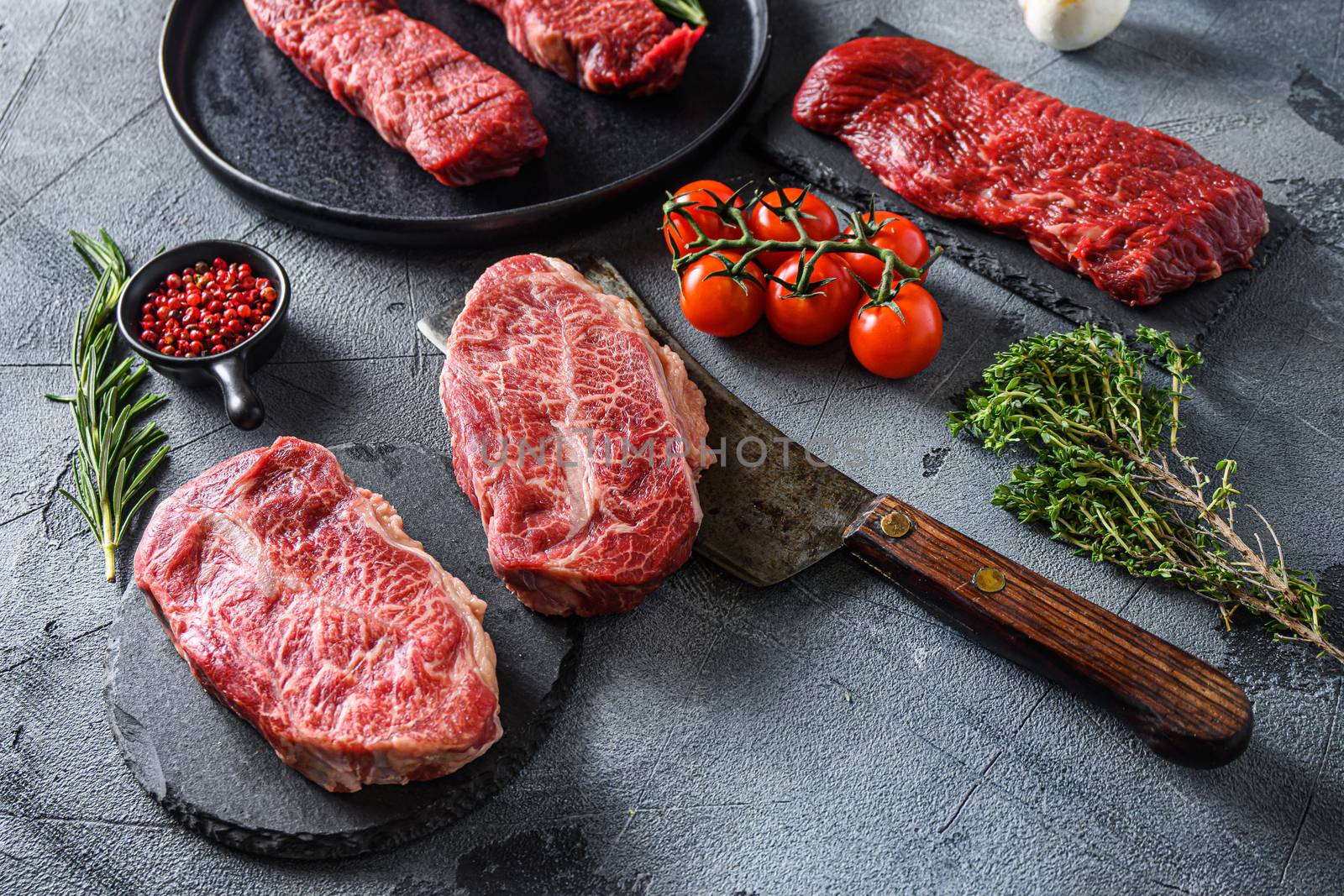 Organic denver, top blade, tri tip steak, machete, skirt steak and , flank, bavette London broil on a black plate and stone slate with seasonings, herbs and meat on butcher vleaver grey concrete background. Side view new wide angle by Ilianesolenyi