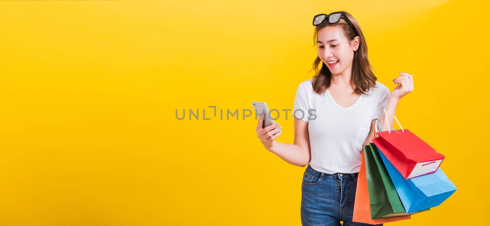 Portrait happy Asian Thai beautiful young woman smile white teeth stand wear t-shirt, She holding shopping bags and using app in mobile phone, studio shot isolated on yellow background with copy space