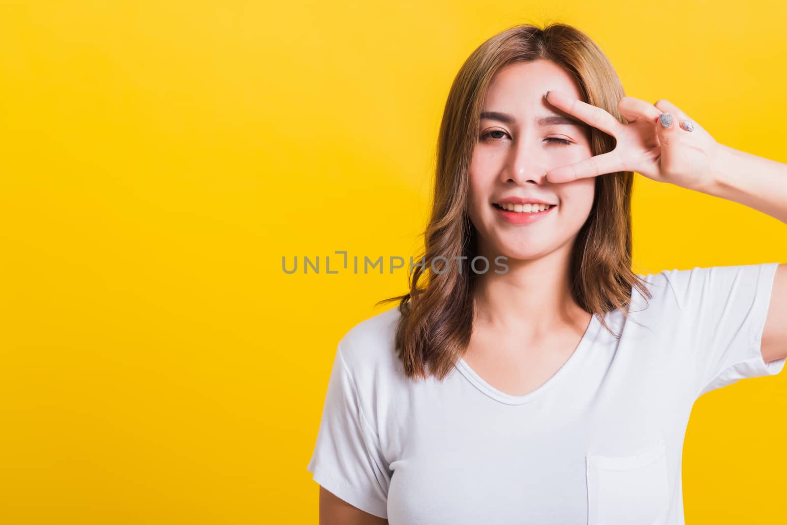 Asian Thai happy portrait beautiful cute young woman smile standing wear t-shirt showing finger making v-sign symbol near eye looking to camera, isolated studio shot yellow background with copy space