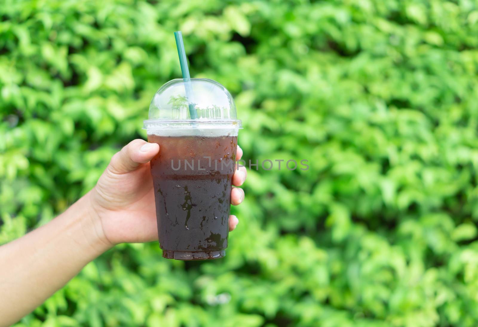 Close up hand holding glass of ice americano coffee with green leaves nature background, selective focus