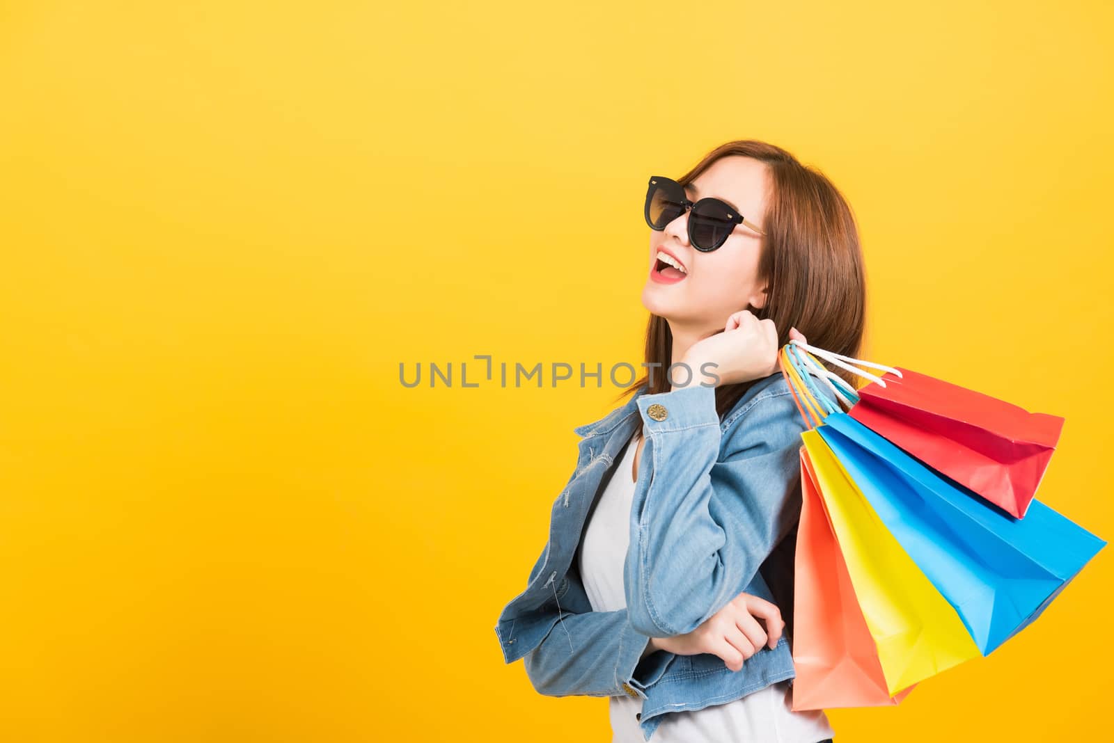 Asian happy portrait beautiful cute young woman teen smiling standing with sunglasses excited holding shopping bags multi color looking up isolated, studio shot yellow background with copy space