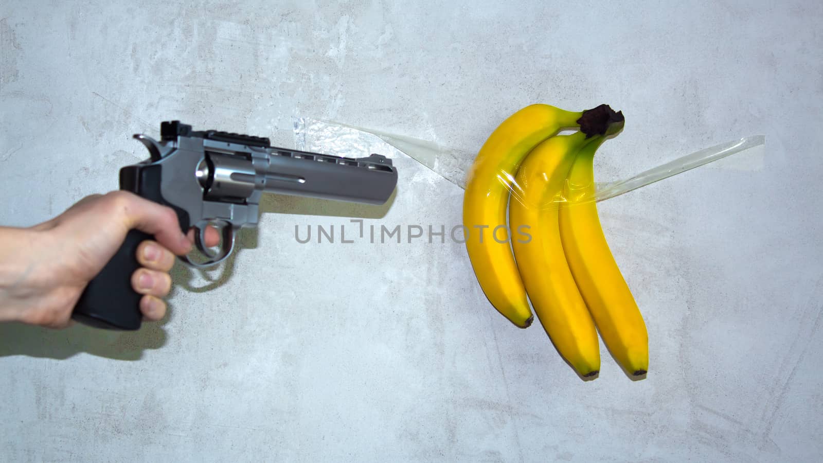 A gun aimed at a banana that is taped to a concrete wall. The concept of weapons and defenselessness. The revolver shoots a banana. Bright yellow bananas hang on the wall on sticky tape.