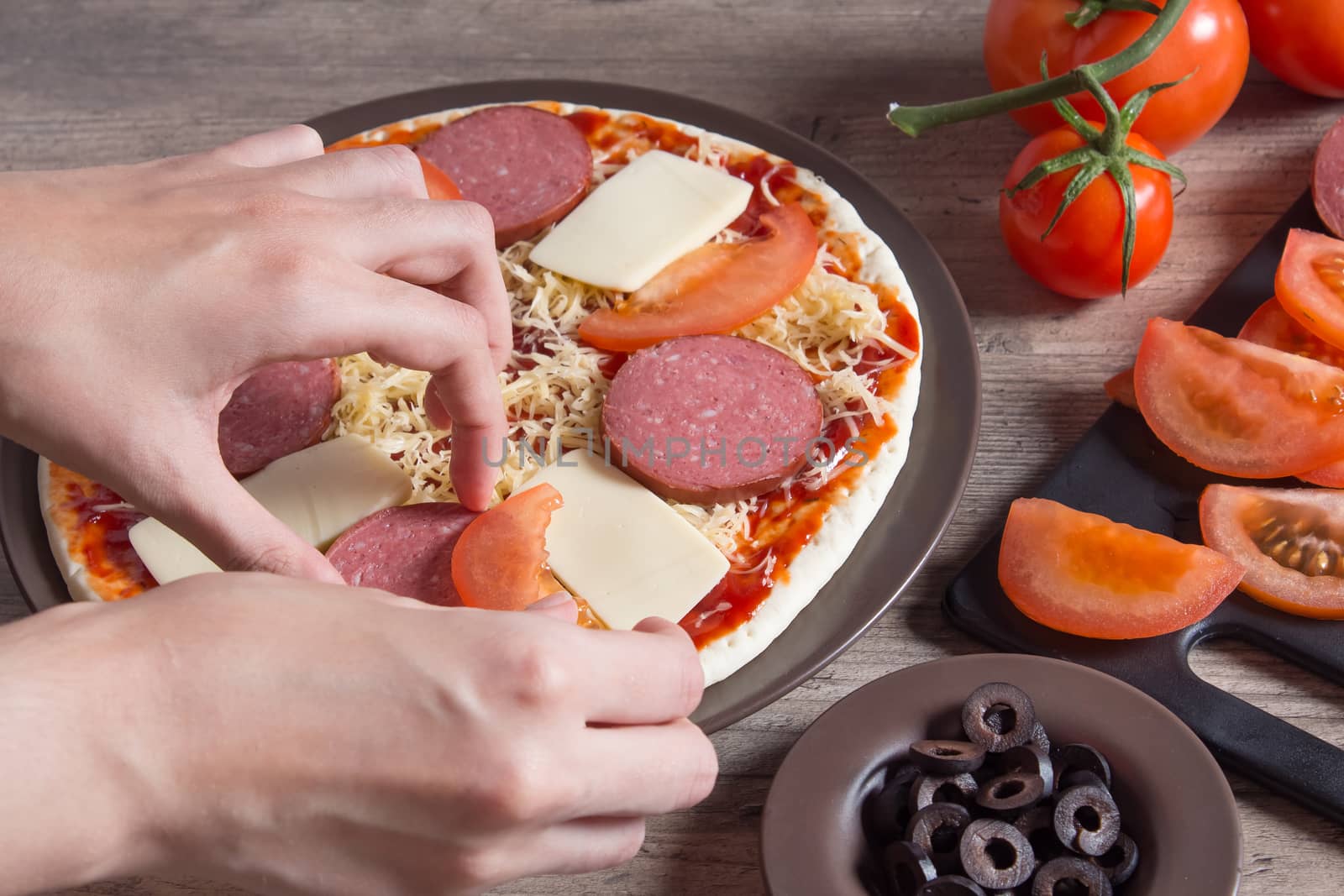 Hands sprinkle the pizza with cheese. On a plate-pizza with tomatoes, olives, bacon, sausage and cheese. The ingredients for making pizza are on a wooden table. Cooking pizza at home.