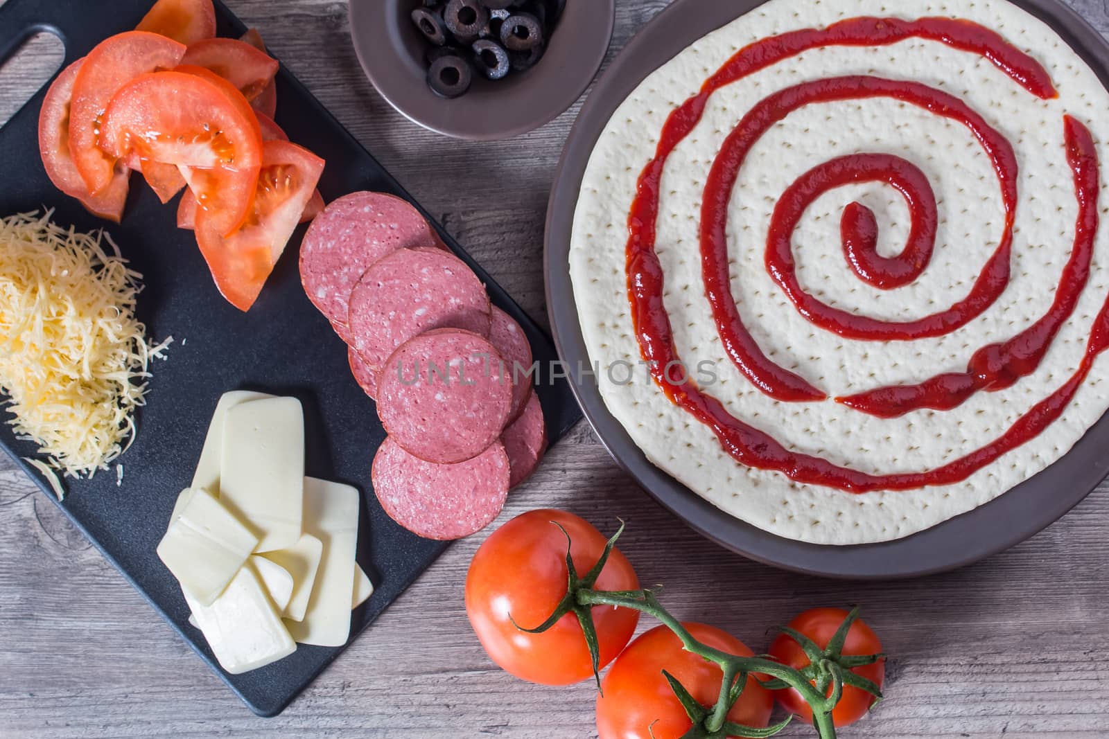 Cooking pizza at home. Pizza base and ingredients for its preparation. Tomatoes, sausage, cheese and olives are on the kitchen table.