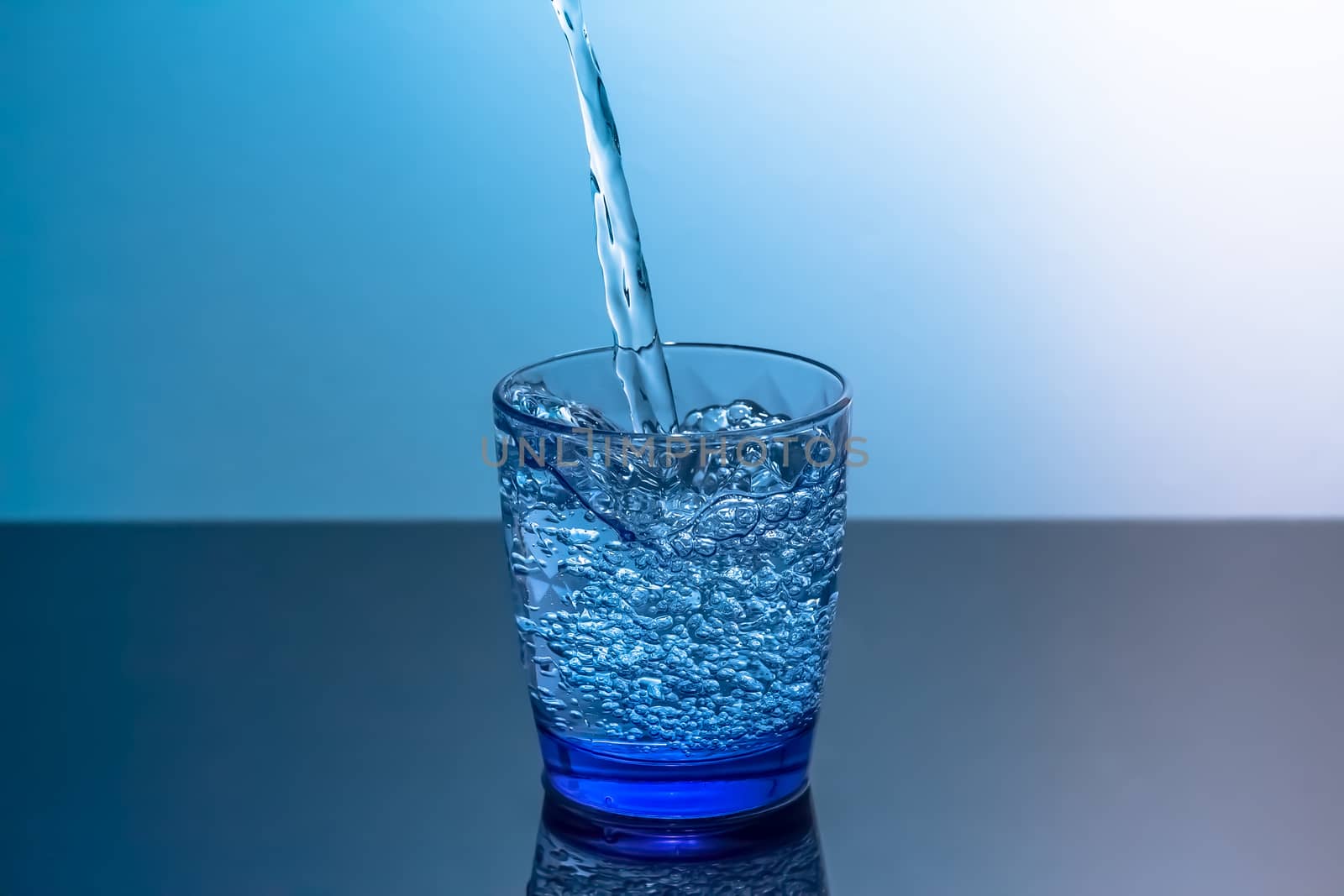 Clean water is poured into a glass standing on a black table. Blue glass with flowing water standing on a black table on a blue background