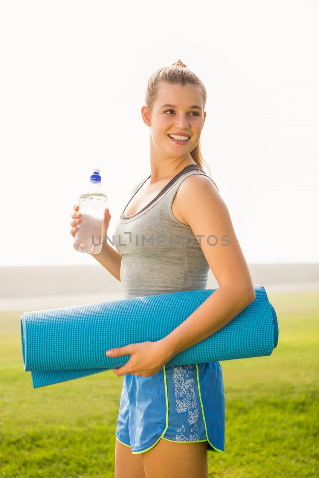 Sporty blonde holding exercise mat and water bottle in parkland