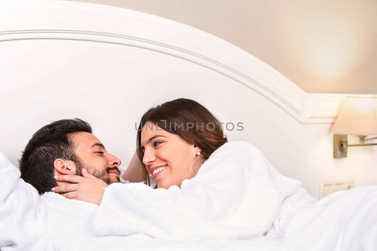 Close up portrait of cute young couple in bathrobe. Laying together on bed in hotel room. Girl showing affection.