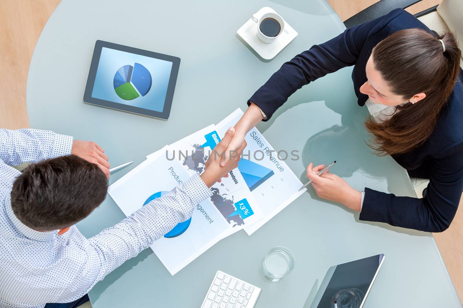 Top view of young business partners shaking hands on deal at desk in office.Documents and digital tablet on table showing statistics and graphics.