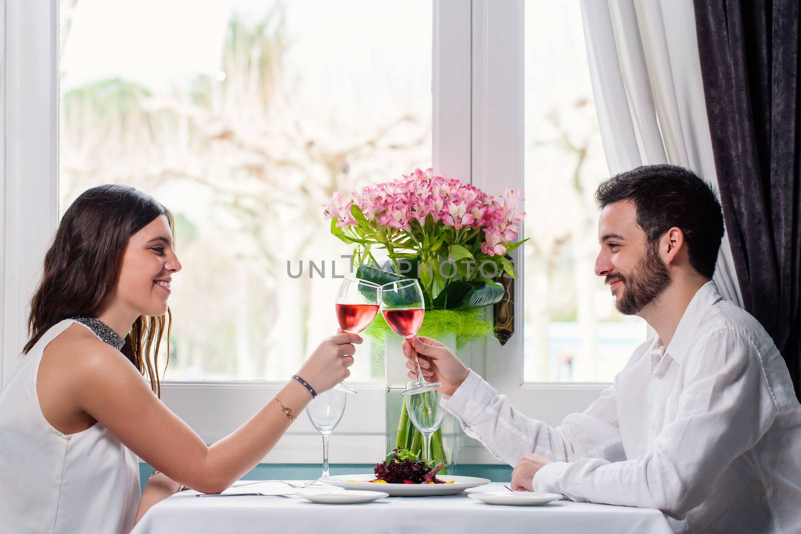 Close up portrait of elegant young couple having romantic dinner in restaurant. Couple sitting next to window with colorful flower bouquet and making a toast with rose wine.