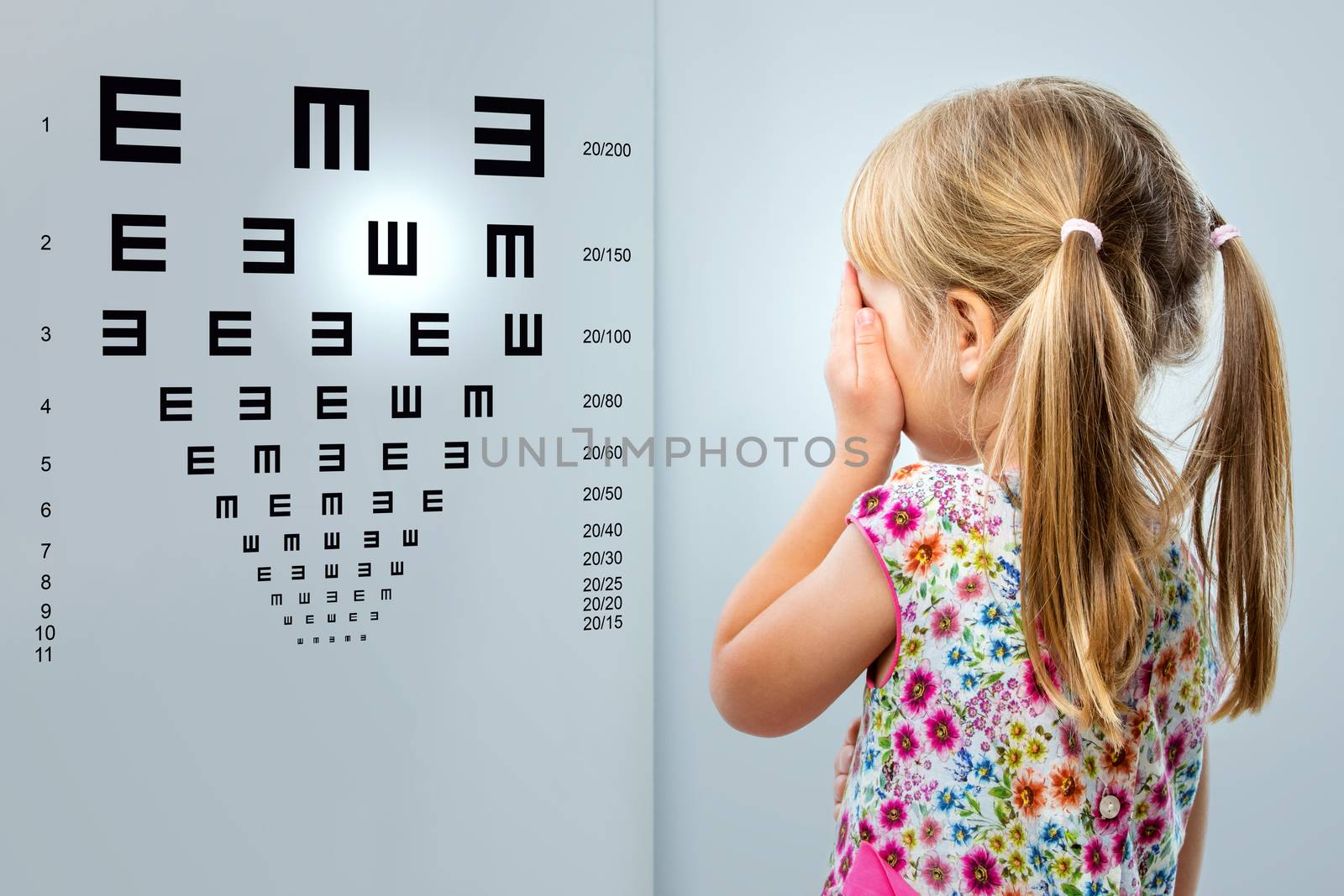 Close up rear view of little girl looking at eye test chart.Kid closing one eye with hand.