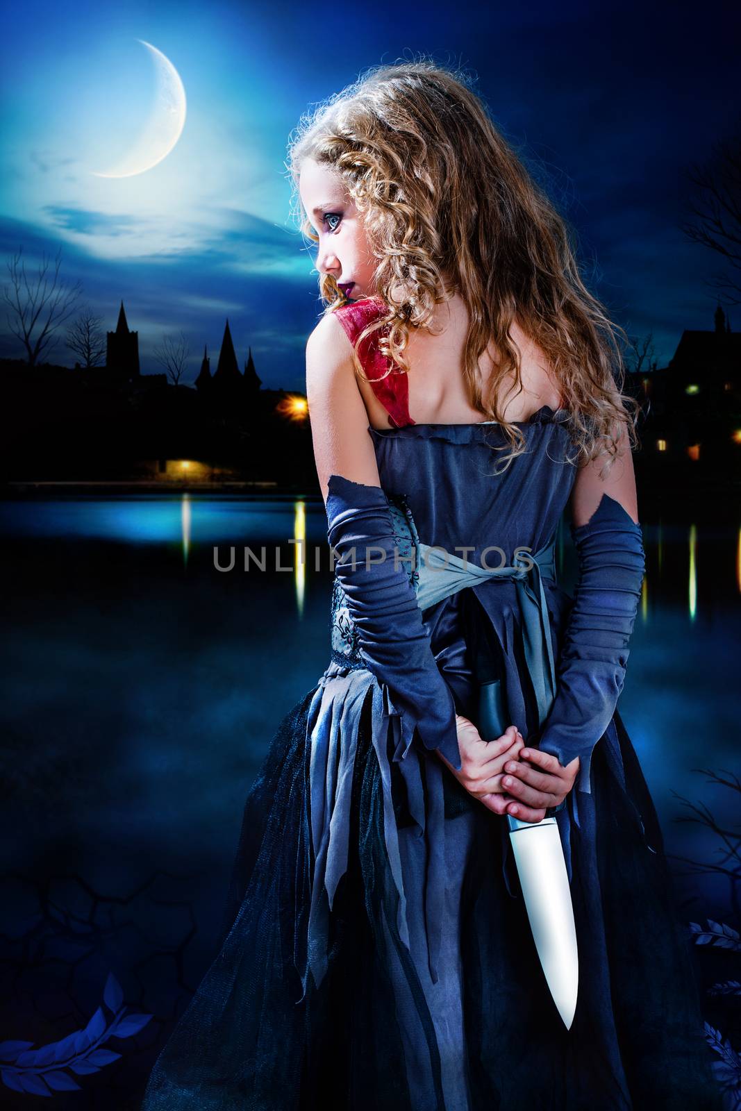 Close up portrait mysterious gothic girl holding knife behind back. Girl standing with big knife behind back at dusk. Medieval cityscape in background with reflection on dark water.