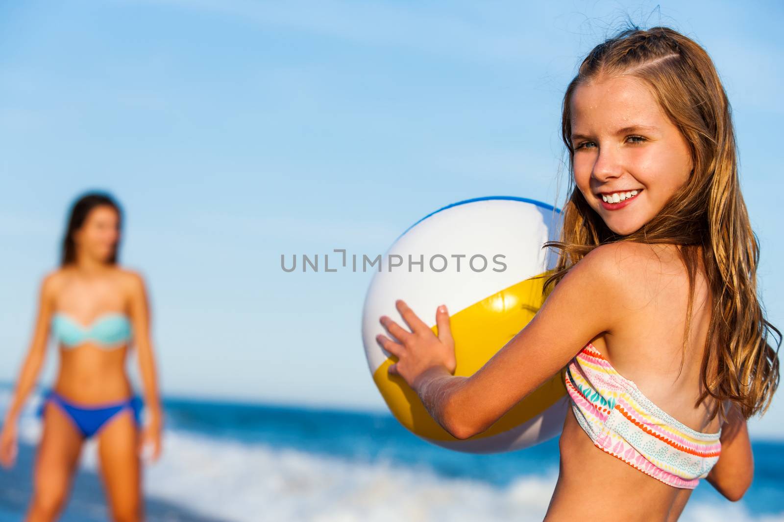 Close up outdoor portrait of young preteen holding inflatable plastic beach ball with mother in background.