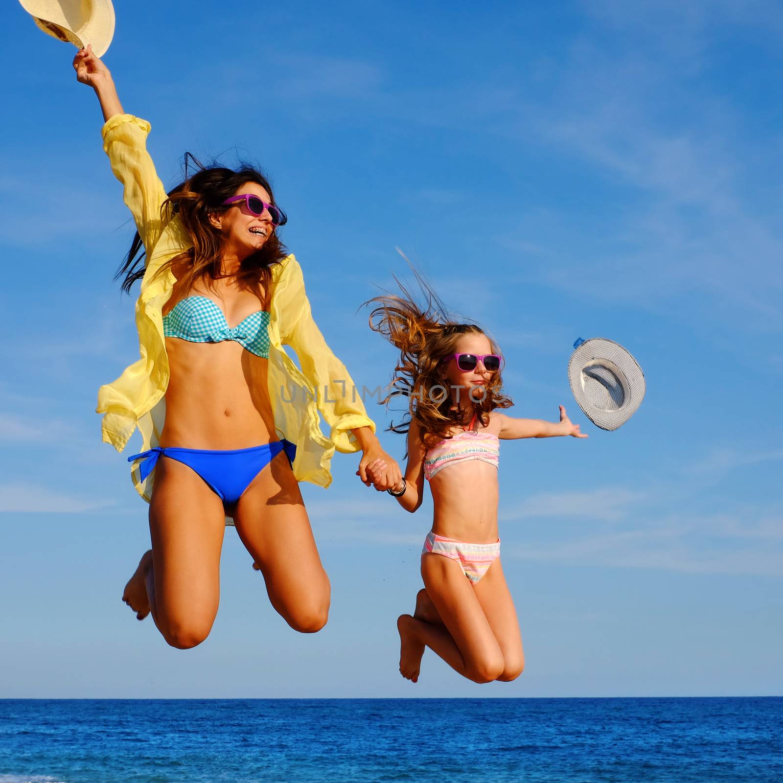 Close up action portrait of young girls on holiday jumping on beach. Two attractive happy women in bikini and sunglasses throwing hats in air.