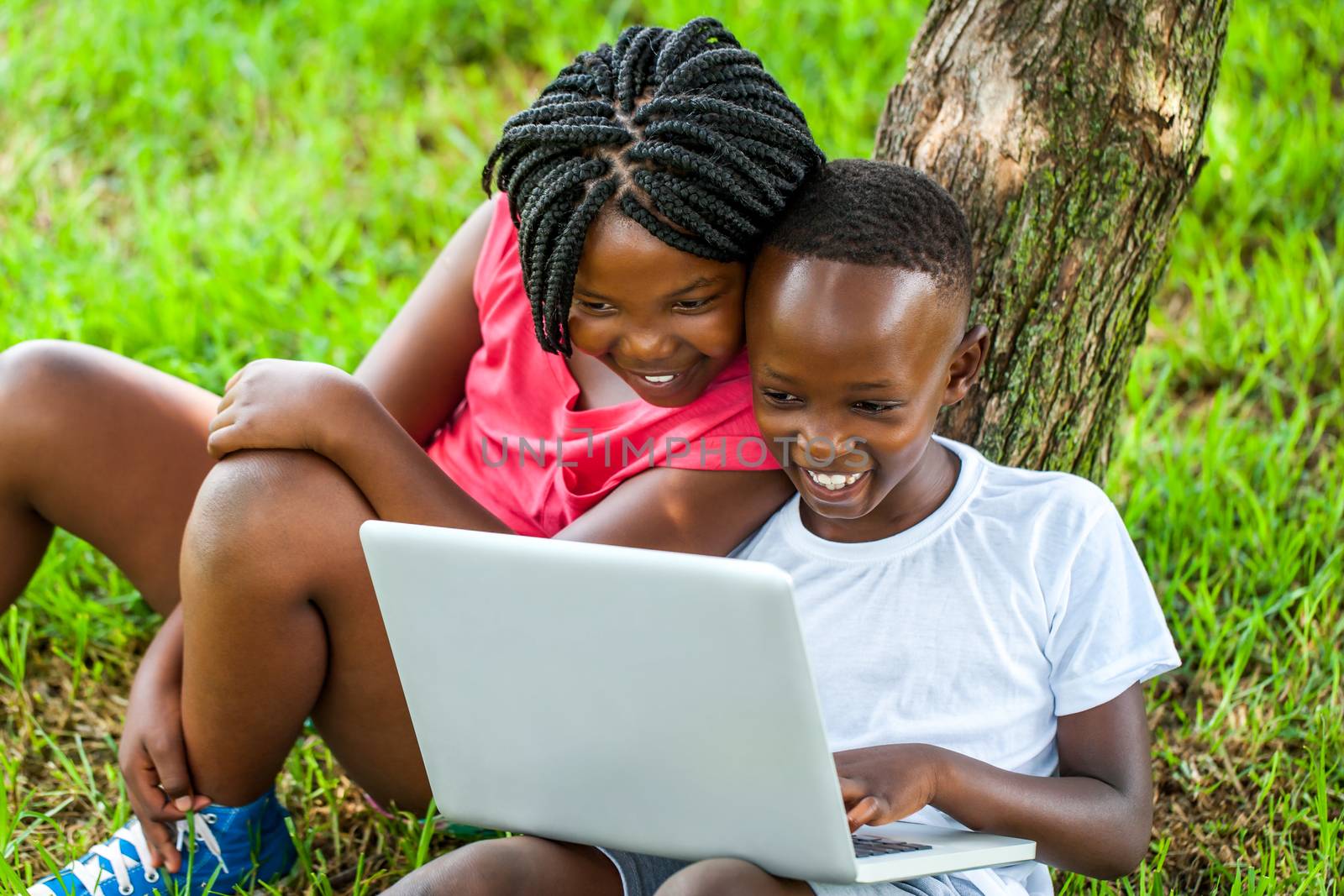 Close up portrait of happy African boy and girl playing on laptop under tree.