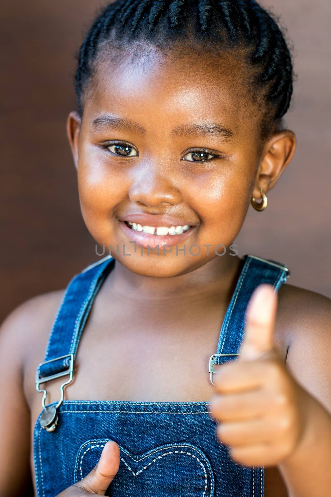 Close up portrait of Cute little african girl against dark brown background doing a thumbs up sign with thumb.