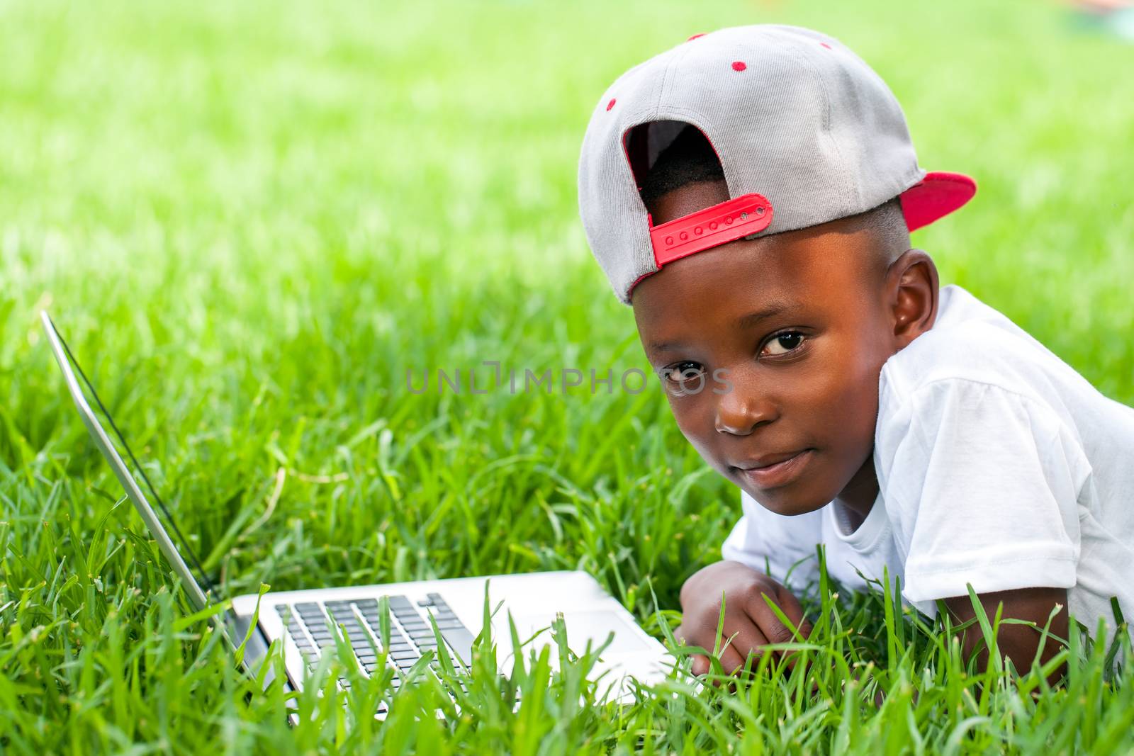 Close up portrait of African boy with baseball hat laying with laptop on grass.
